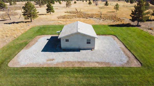 A house surrounded by cleared gravel and low green landscaping with dry brush and trees in the distance